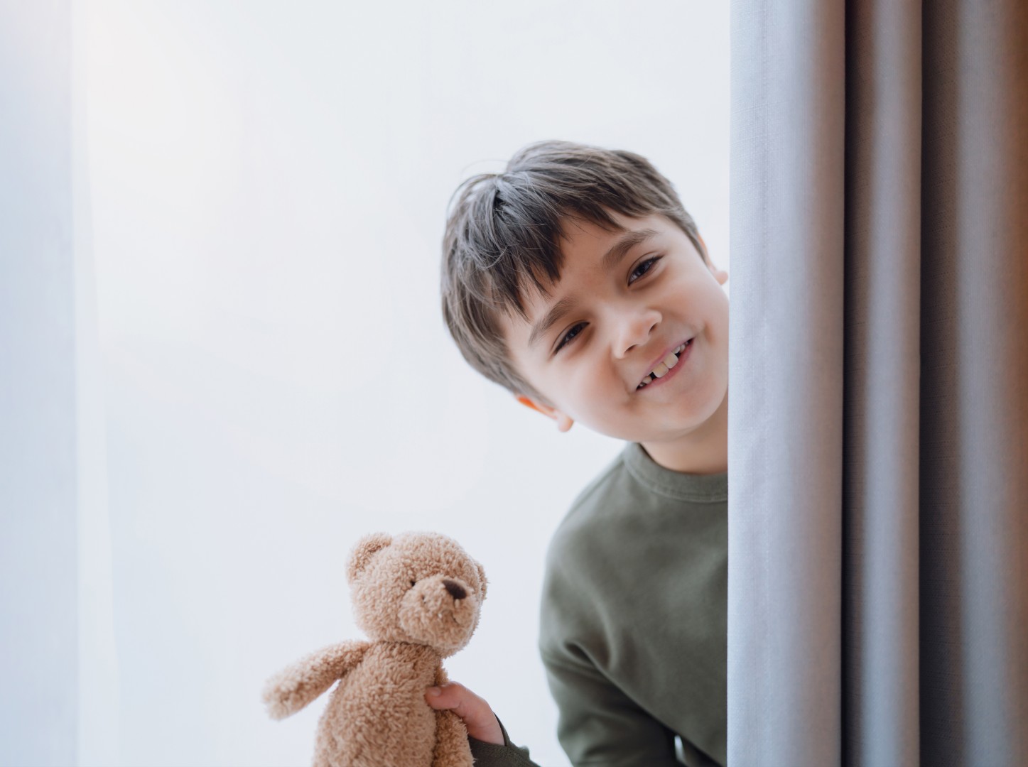 Cute young boy holding teddy bear standing behind lace curtain with bright light in morning, Happy kid with smiling face on window sill and plays at hide-and-seek behind curtain